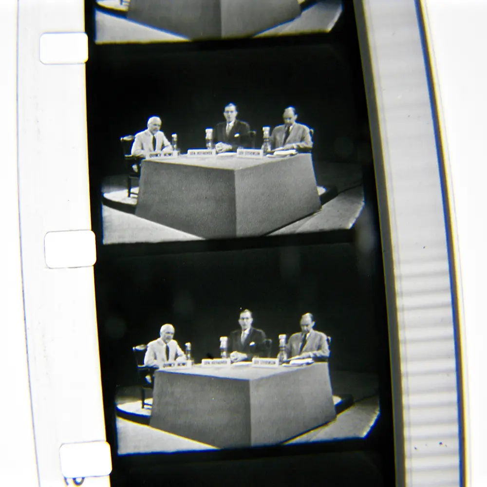 An image of several frames from a film. The film shows three men in suits seated at a table with microphones.