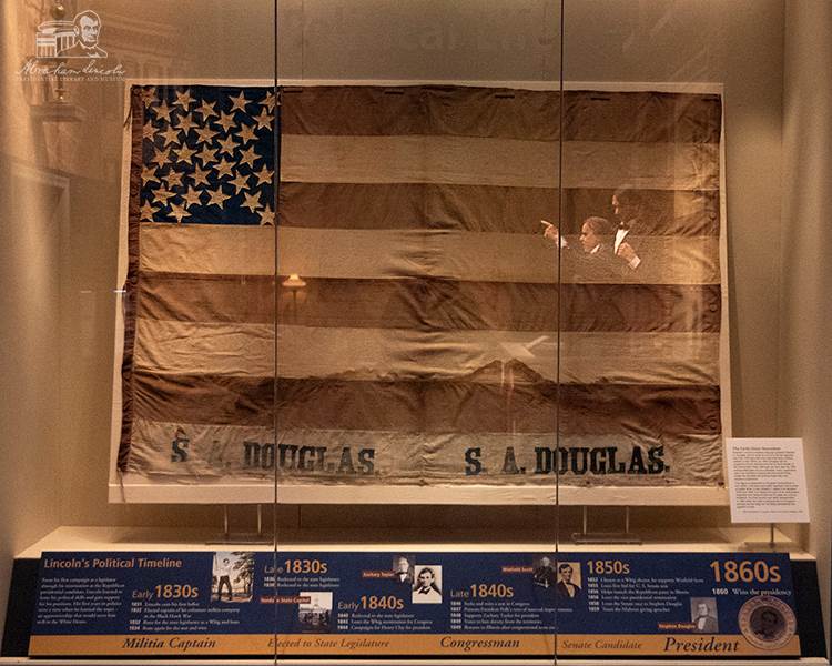 The banner in its display case, where a reflection from an exhibit showing the Lincoln-Douglas debates can be seen.